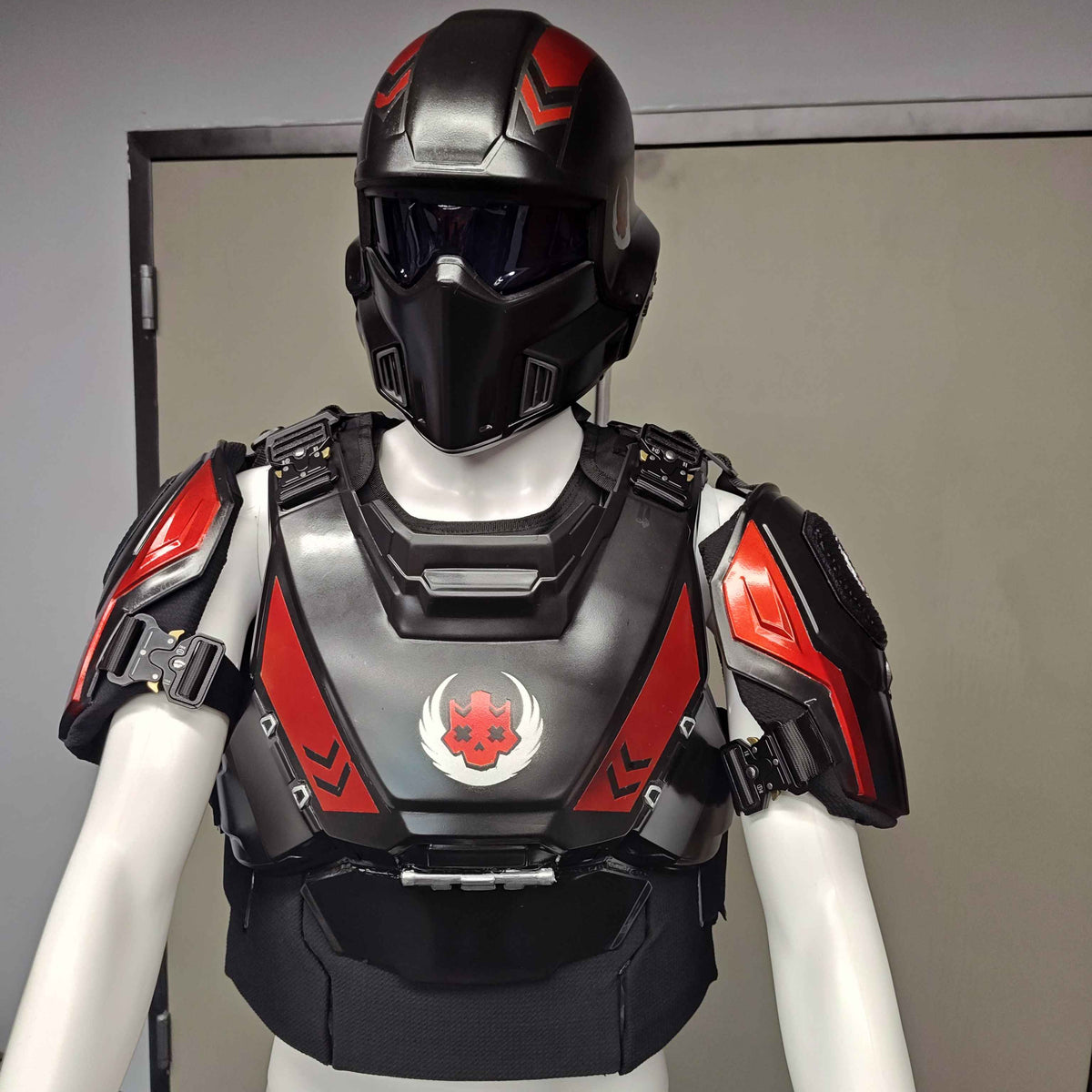A black and red futuristic armored chest plate with matching helmet displayed on a white mannequin torso in an indoor room.