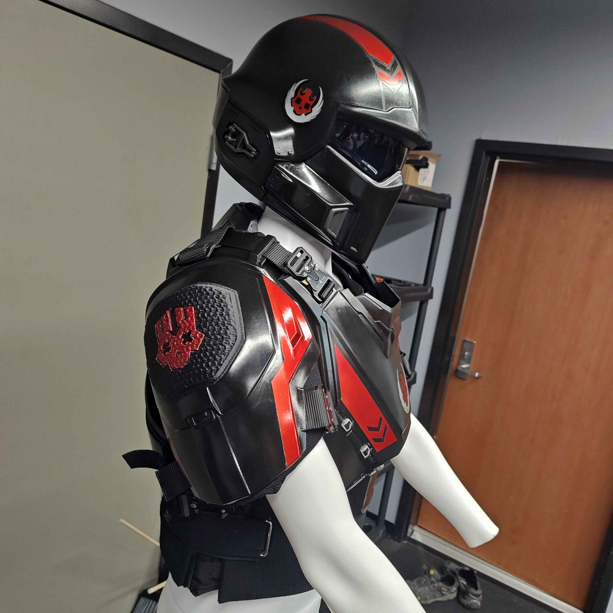 A black and red futuristic armored chest plate and matching helmet displayed on a white mannequin torso in an indoor room with a door and shelving visible.