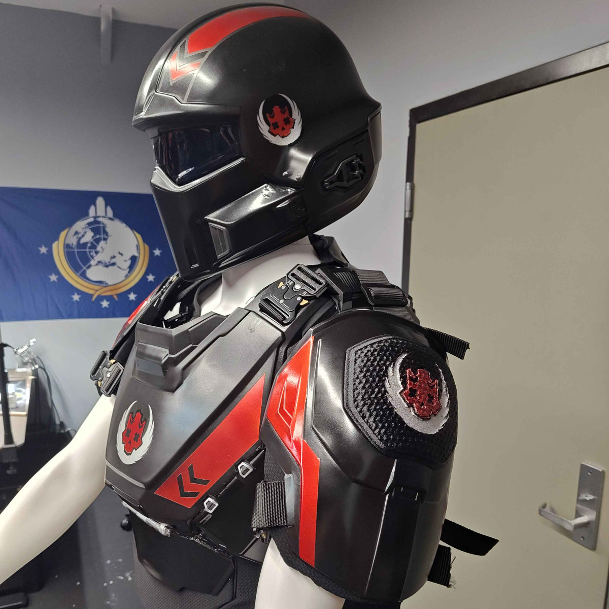 A black and red sci-fi style helmet and matching armored chest rig displayed on a white mannequin torso in an indoor room with a door and wall banner visible in the background.